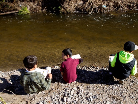 3 children sitting on the edge of the Santa Cruz River with paper in their hands